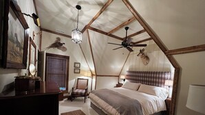 A cozy, rustic bedroom with a large bed, wooden beams, a ceiling fan, and a mounted animal head on the wall. - Twin River Stone Cottage - 7 BR/Sleeps 20 (Norfork)
