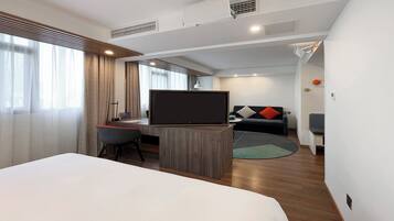 A modern hotel room with a large bed, a desk with a flat-screen TV, a sofa, and a small rug.