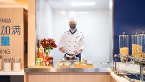 Ein Koch in weißer Uniform und Kochmütze steht hinter einer Theke mit verschiedenen Gewürzen, einer Blumenvase und Lebensmitteln.