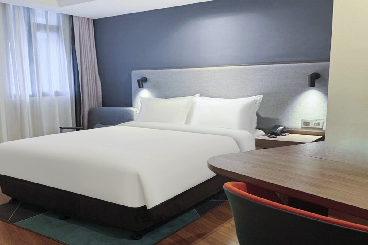 A hotel room with a bed, a desk, a chair, a nightstand, and a lamp.