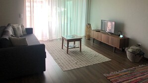 A living room with a sofa, a coffee table, a TV on a wooden stand, and a window with sheer curtains. - Apartment with view and pool access 900 meters from the beach (Ovar)