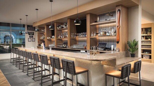 Zona de bar moderna con una barra larga, varios taburetes y estanterías llenas de botellas.