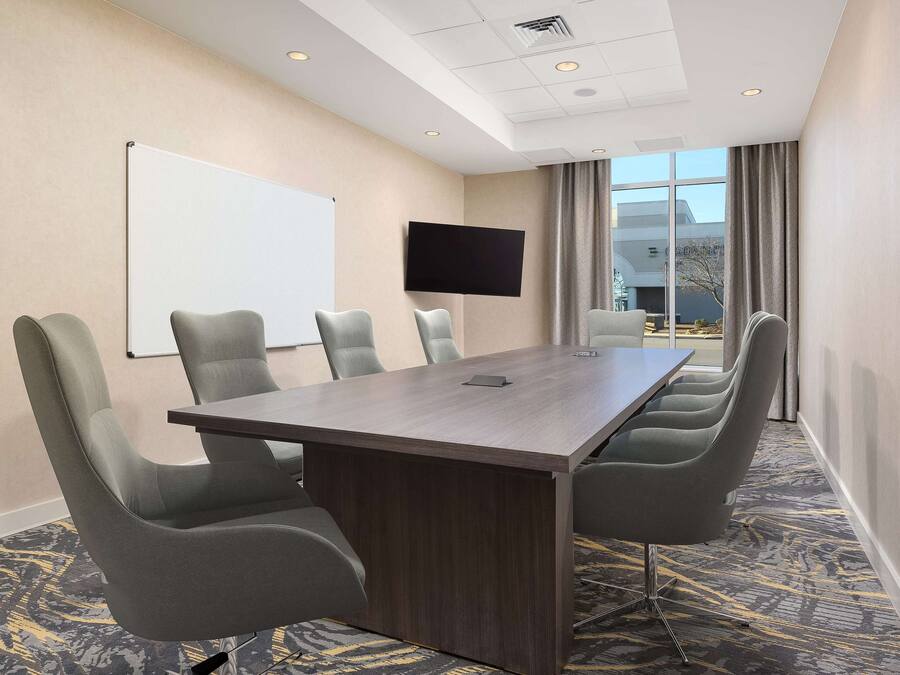 Una sala de conferencias con una mesa grande, seis sillas, una pizarra, un televisor de pantalla plana y ventanas amplias con cortinas.
