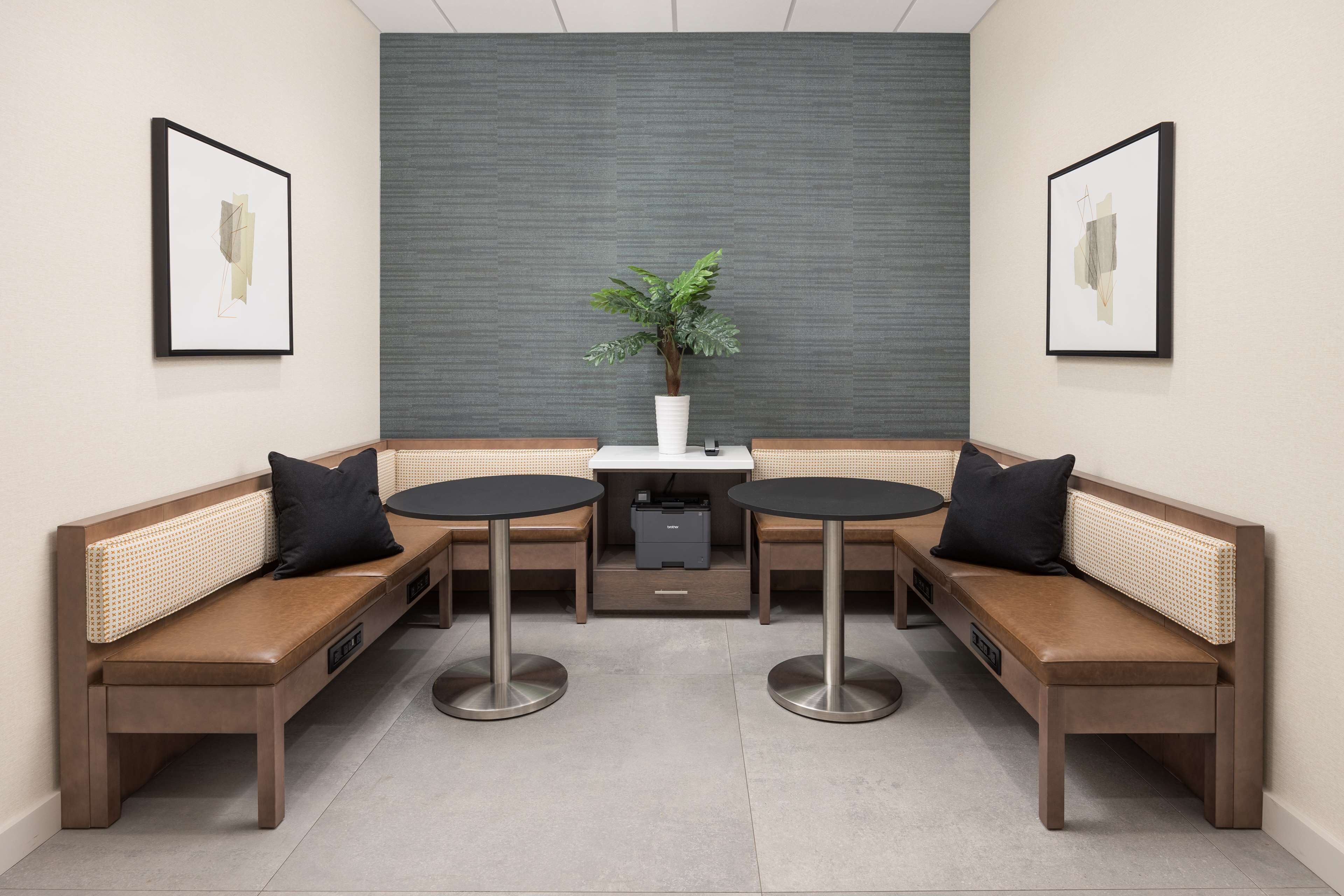 A modern seating area with two round tables and benches, a central potted plant, and framed artwork on the walls.