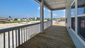 Een overdekt houten terras met een hekwerk, met uitzicht op een woongebied met huizen en een strand.