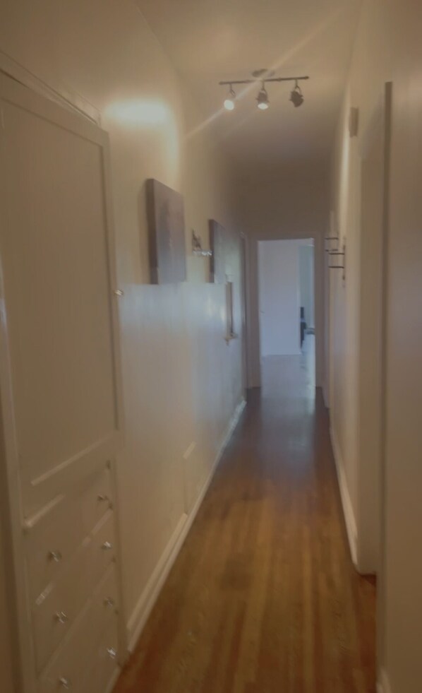 A narrow hallway with wooden flooring and white walls. There are doors on the left and right, and a light fixture on the ceiling.
