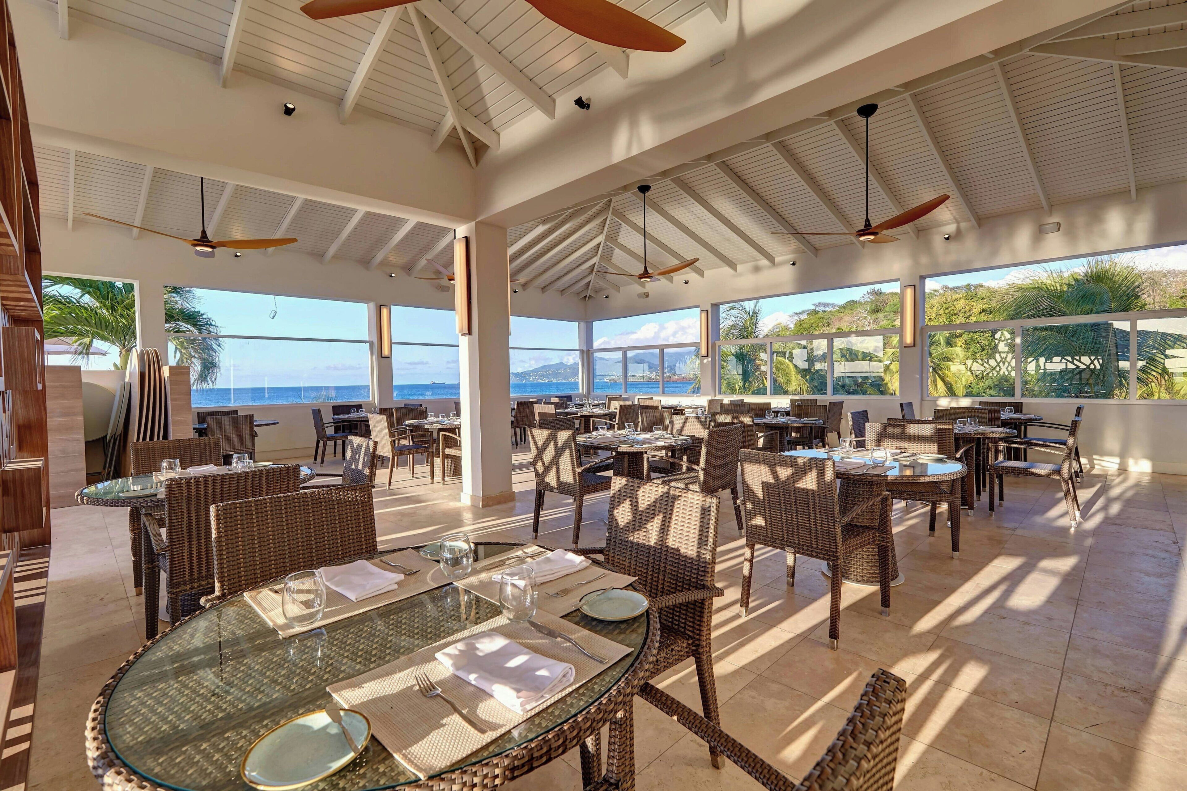 Un área de comedor al aire libre amplia, con mobiliario de mimbre, mesas de vidrio y ventiladores de techo.