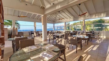 Un área de comedor al aire libre amplia, con mobiliario de mimbre, mesas de vidrio y ventiladores de techo.
