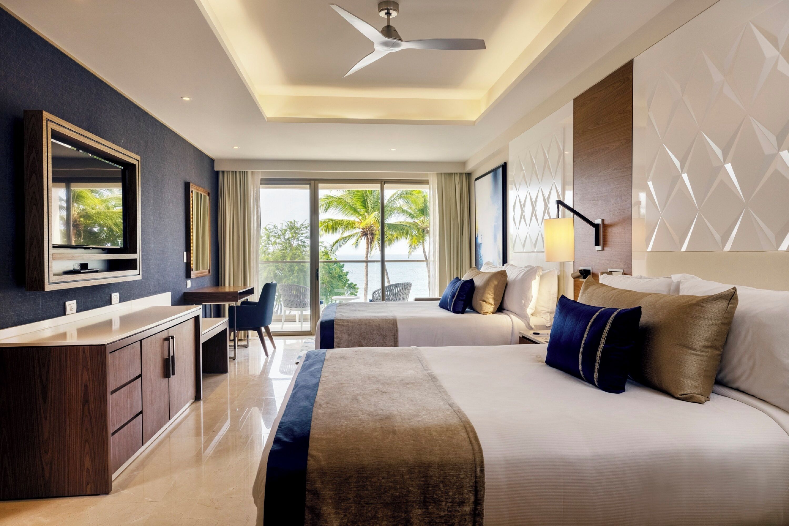 A hotel room with a bed, a desk, a chair, a TV, and a view of palm trees. - Royalton Grenada (St. George's)