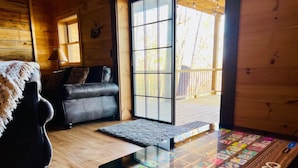 A wooden cabin interior with a sofa, a glass-top coffee table displaying colorful items, and a large window offering a view of the outdoors. - Stunning Views over Aska +Hot Tub & Firepit +Arcade and Pool table +Near Toccoa (Blue Ridge)