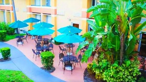 Un espace extérieur aménagé en terrasse, avec des tables et des chaises, entouré d’une végétation luxuriante et bordé d’un bâtiment à la façade jaune.