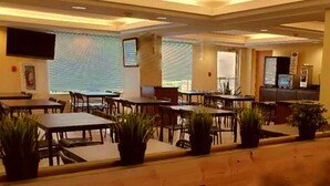 Une salle à manger d’hôtel avec plusieurs tables et chaises, des plantes en pot et un écran de télévision fixé au mur.