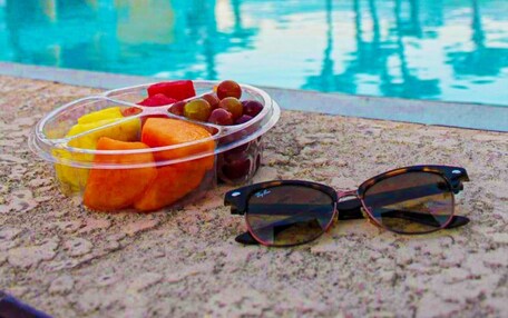 A swimming pool with a clear view of the poolside area, a fruit bowl with various fruits, and a pair of sunglasses on the edge of the pool.