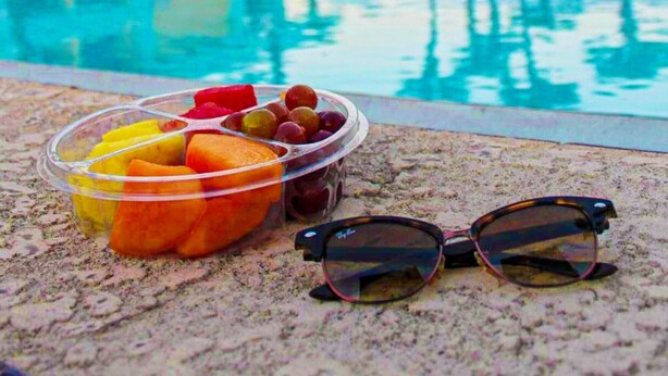 Une piscine offrant une vue dégagée sur l’espace qui l’entoure, un panier de fruits rempli de différentes sortes de fruits, et une paire de lunettes de soleil posée sur le bord de la piscine.