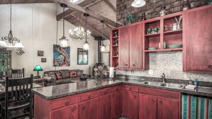 A spacious kitchen with red cabinets, a granite countertop, and a large island. There is a dining area with a wooden table and chairs, and a cozy seating area with a sofa and a lamp. - A Climbing Cub by Ruidoso Vacation Rentals (Ruidoso)