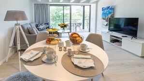 Un soggiorno moderno con un tavolo da pranzo apparecchiato per la colazione, una TV e un balcone con vista sugli alberi.