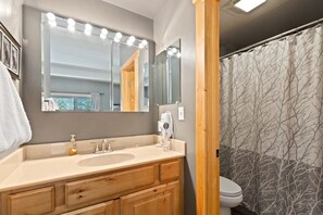 A bathroom with a vanity, sink, and mirror with lights. There is a shower curtain with a tree pattern.