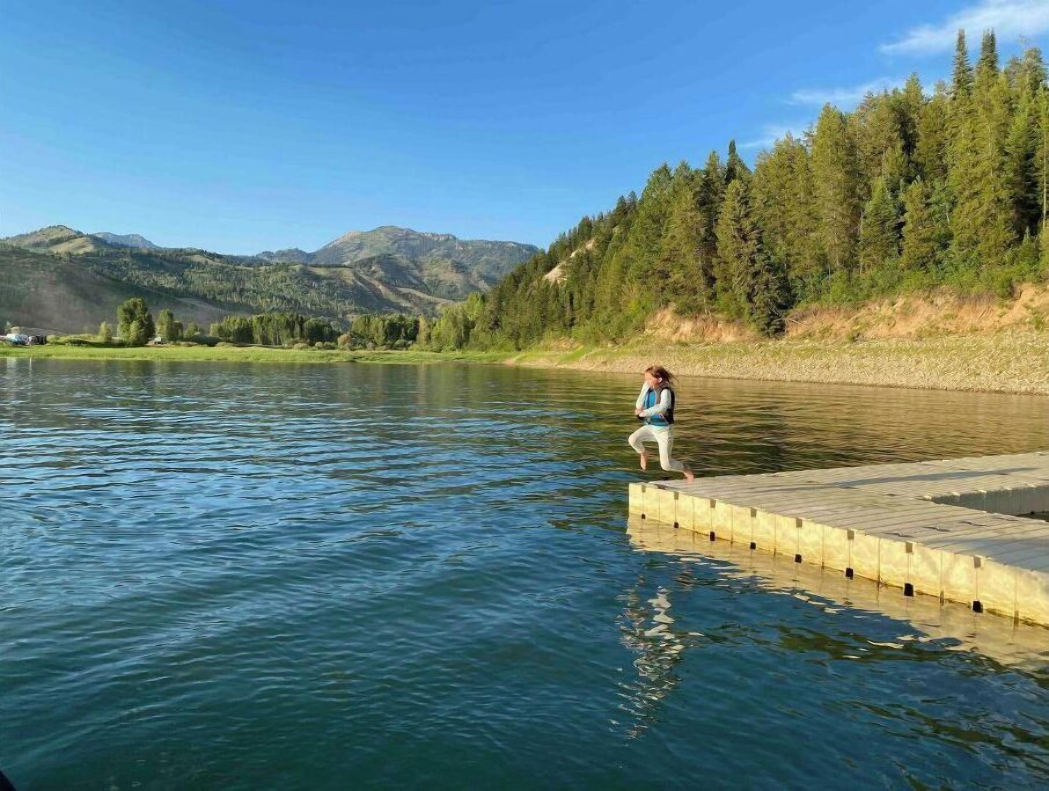 Eine Person steht auf einer hölzernen Anlegestelle, die sich in einen ruhigen Fluss erstreckt, umgeben von üppigem Grün und Bergen im Hintergrund.