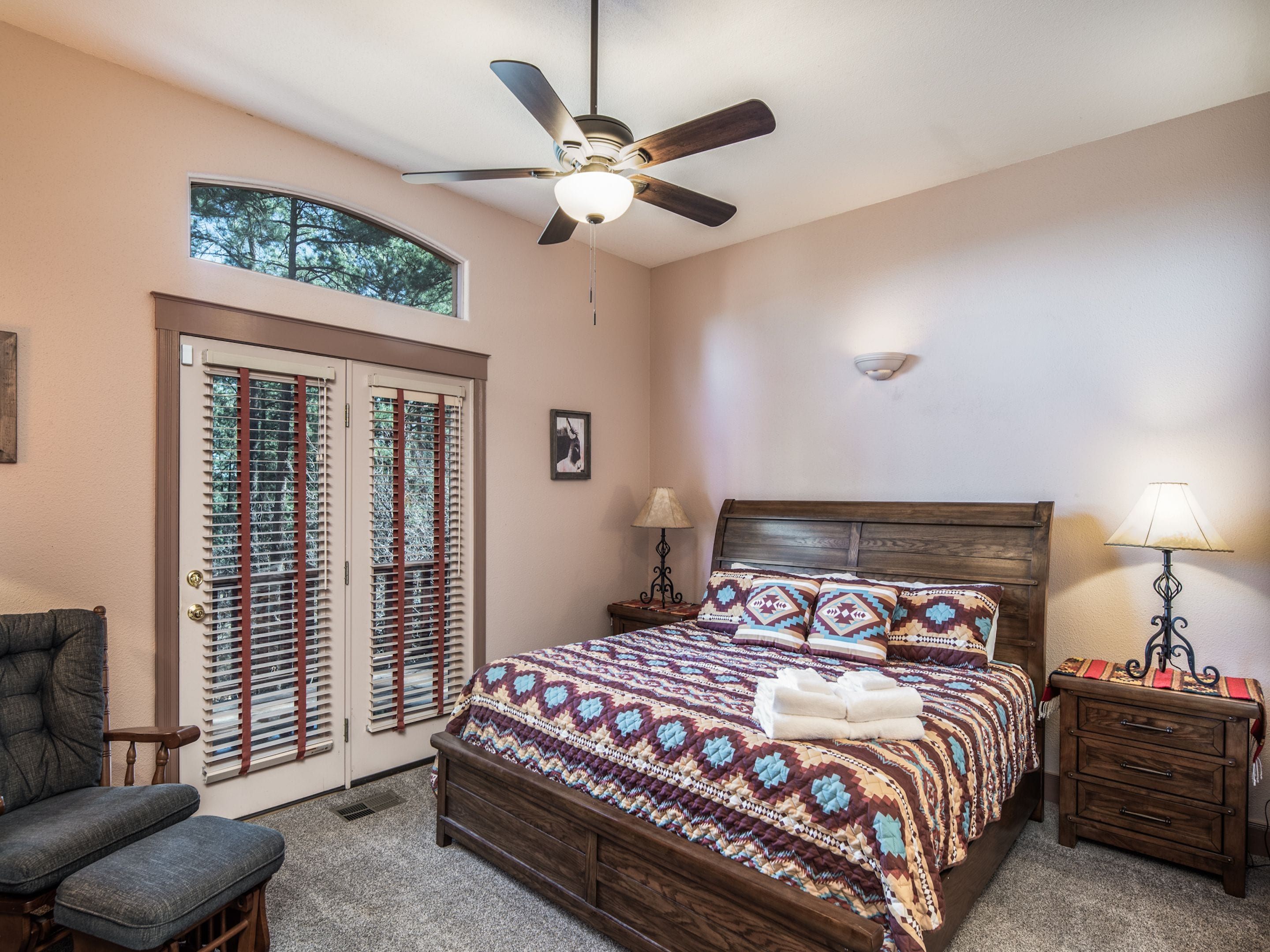 A bedroom with a wooden bed, a ceiling fan, a window with blinds, a chair, and a nightstand with a lamp.