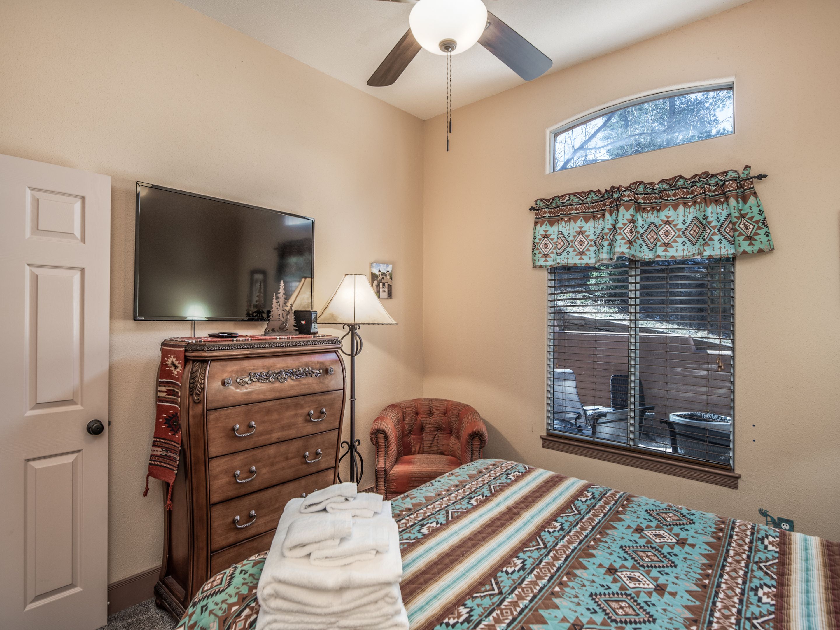 A bedroom with a bed, a dresser, a chair, a television, a window with blinds, and a ceiling fan.