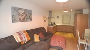 Una sala de estar con un sofá marrón, una mesa de comedor con sillas, una zona de cocina y un cuadro de Buda en la pared.