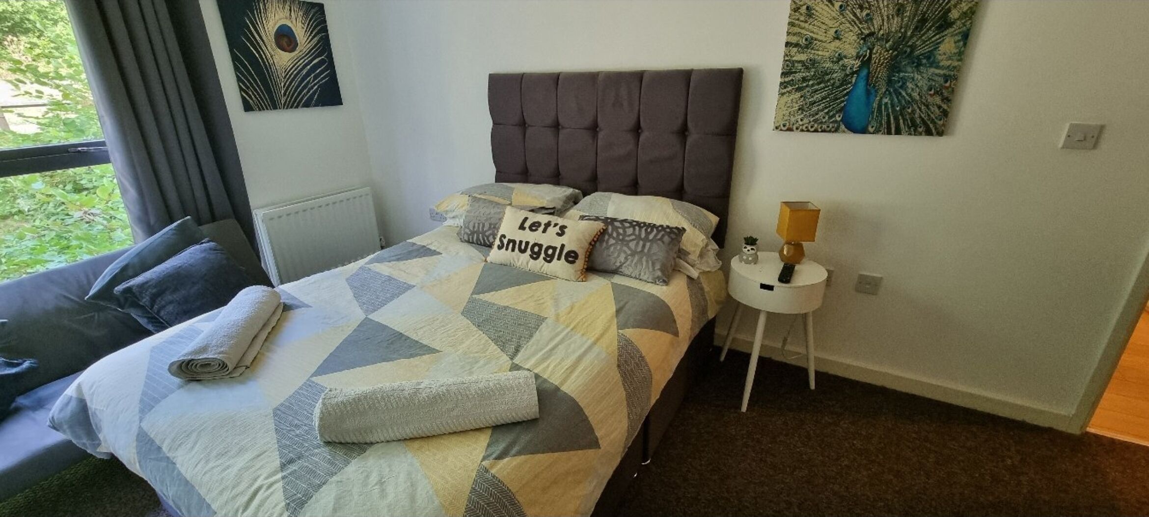 A neatly made bed with a geometric patterned bedspread, a pillow with the text "Let's Snuggle," and a rolled towel on the bed.