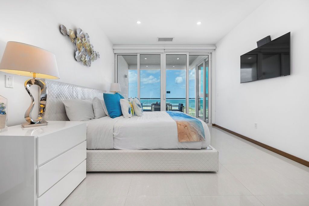 Une chambre moderne avec un lit, une table de chevet, une lampe et une vue sur l’océan grâce à de grandes portes vitrées.