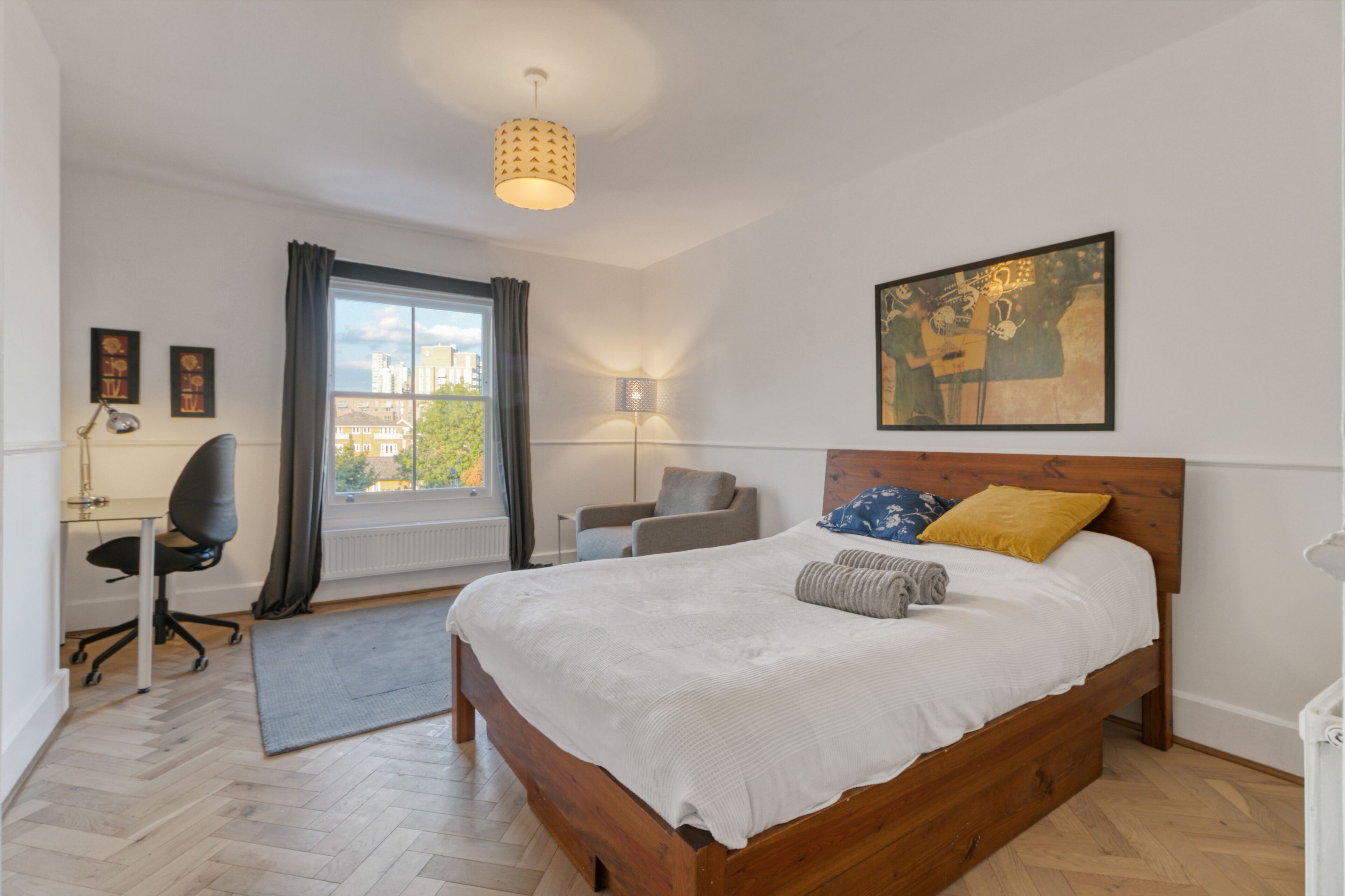 A bedroom with a wooden bed, a desk with a chair, a window with curtains, and a city view.
