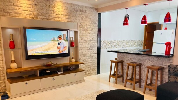 A modern living room with a flat-screen TV, a wooden entertainment unit, a black sofa, and a kitchen area with stools and red pendant lights. - Modern and cozy house with fireplace and heated pool just 50m from the beach. (Imperador - Praia Grande)