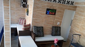 A modern lounge area with a sofa, chairs, and a small stool. A neon sign indicates a bar is nearby. - Modern and cozy house with fireplace and heated pool just 50m from the beach. (Imperador - Praia Grande)
