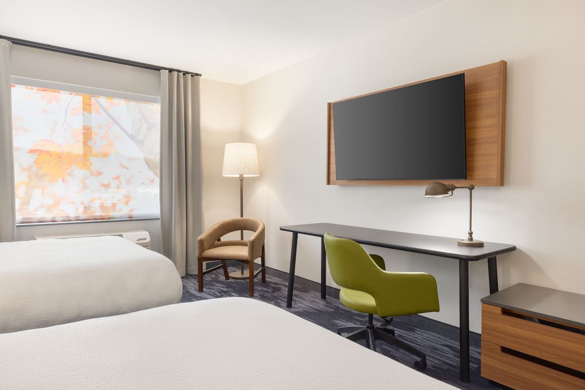 A hotel room with two beds, a desk, a chair, a lamp, and a large window with a view of autumn leaves.