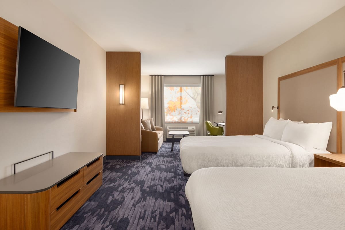 A hotel room with two beds, a flat-screen TV, a desk, a chair, and a window with curtains.