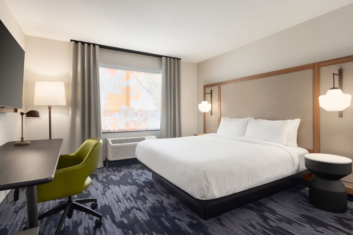 A hotel room with a bed, a desk, a chair, a lamp, and a window with curtains.