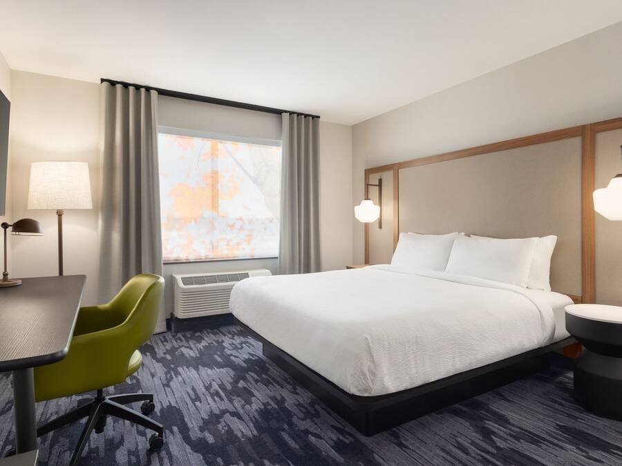 A hotel room with a bed, a desk, a chair, a lamp, and a window with curtains.