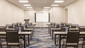 A conference room with rows of tables and chairs, a projector screen, and water bottles on each table.