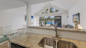 Una cocina con encimera de granito, pileta de acero inoxidable y salpicadero de azulejos blancos. Hay un comedor con mesa de vidrio y sillas blancas.