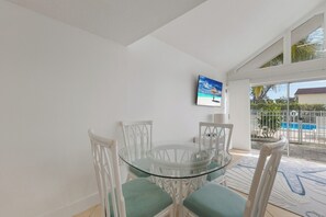 Un comedor con mesa de vidrio y sillas blancas, un cuadro de una playa y vistas a una piscina exterior.