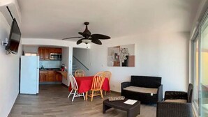 Un salon avec un canapé, des fauteuils, une table basse, un téléviseur fixé au mur et une cuisine en arrière-plan.