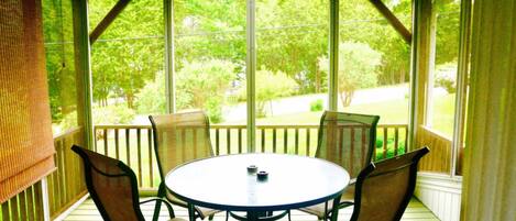 A covered outdoor seating area with a round table and four chairs, large windows, and a view of greenery.