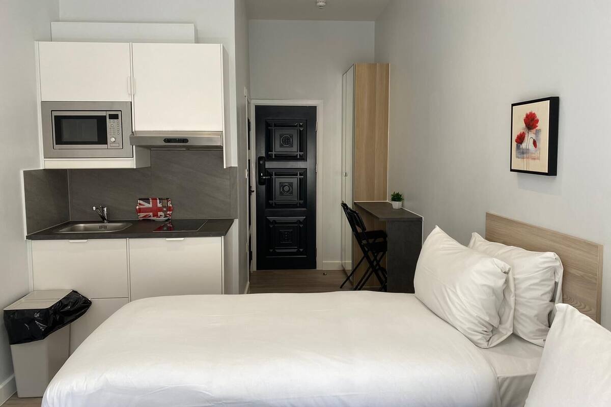 A compact hotel room with a bed, a kitchenette, a small desk, and a wall-mounted picture.