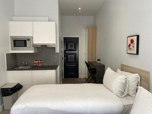 A compact hotel room with a bed, a kitchenette, a small desk, and a wall-mounted picture. - MSK Premium (London)