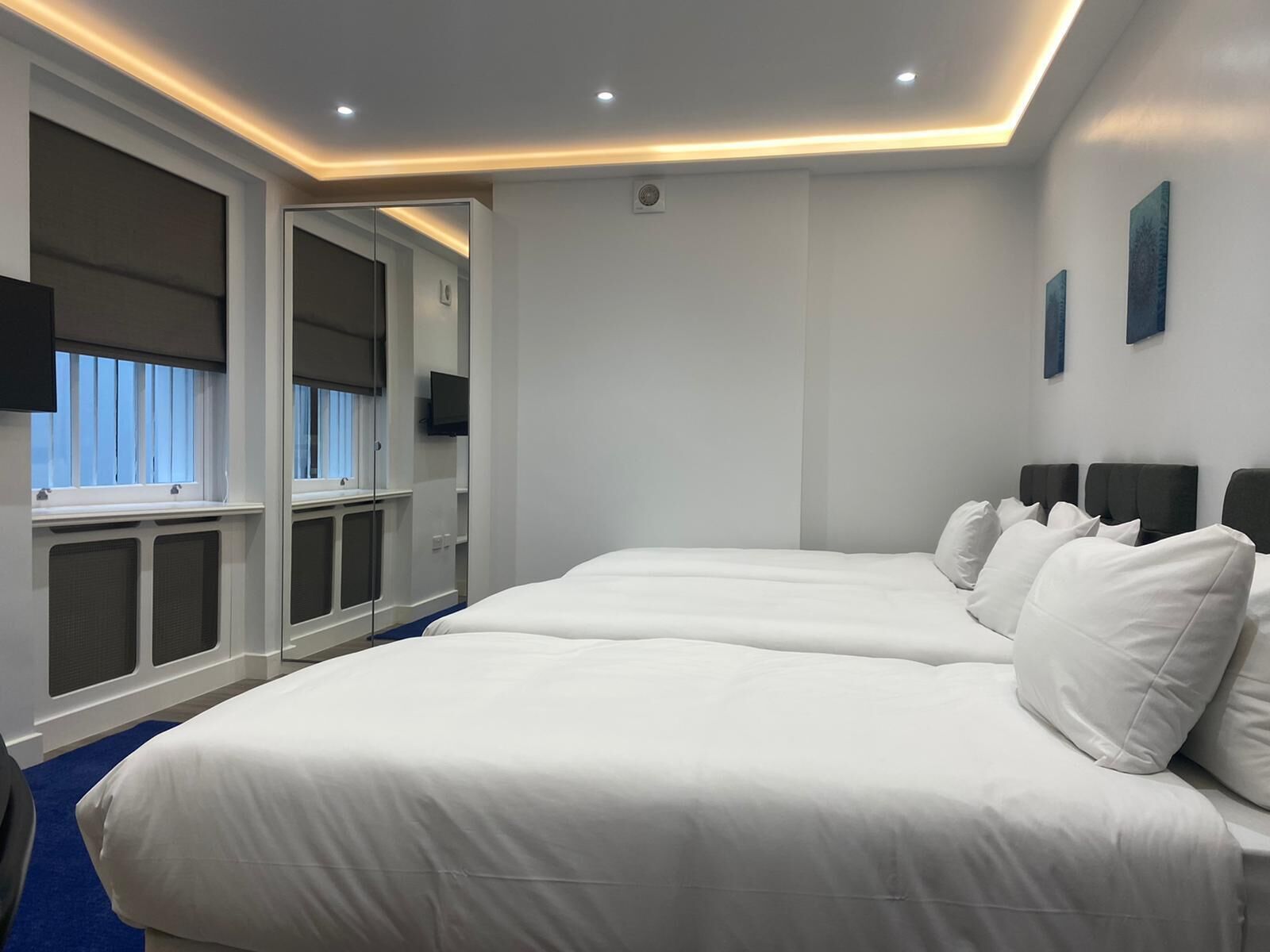 A modern hotel room with two beds, a large mirror, a TV, and a window with blinds.