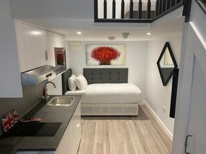 A compact hotel room with a kitchenette, a bed with a grey headboard, a red floral artwork on the wall, and a black-framed mirror. - MSK Premium (London)