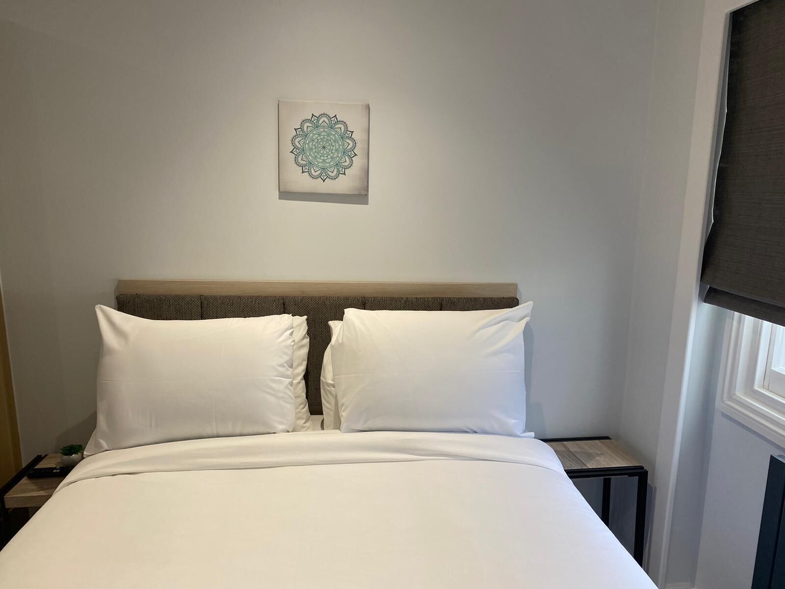 Ein ordentlich bezogenes Bett mit weißen Leinen und einem Kopfteil, ein kleiner Nachttisch mit Lampe sowie ein an der Wand befestigtes dekoratives Element.