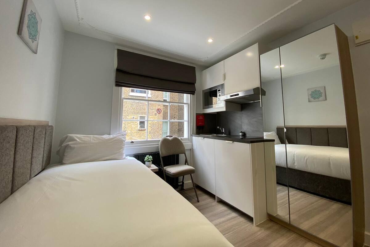 A compact hotel room with a bed, a small kitchenette, a chair, and a large mirror.