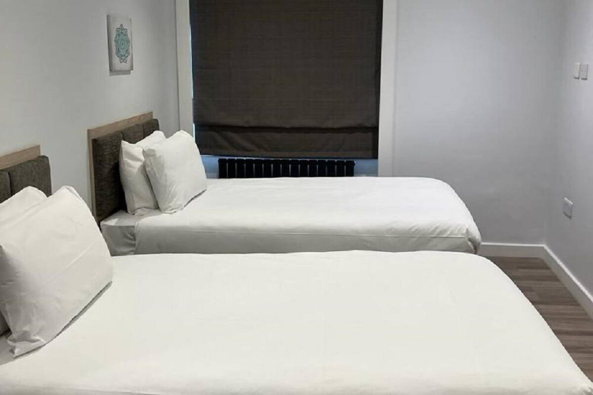 A room with two beds, a large window with a dark blind, and a ceiling with recessed lighting.
