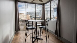 Ein Eckzimmer mit rundem Tisch und vier Stühlen, großen Fenstern mit Vorhängen, einem Deckenventilator und Blick auf Stadtgebäude.