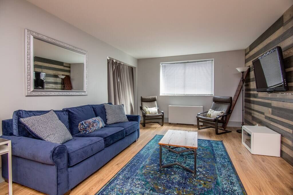 A living room with a blue sofa, two armchairs, a coffee table, a large mirror, a window with blinds, and a radiator.