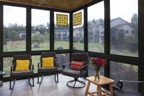 A room with three rocking chairs, a small table with a vase of flowers, and large windows offering a view of houses and trees.