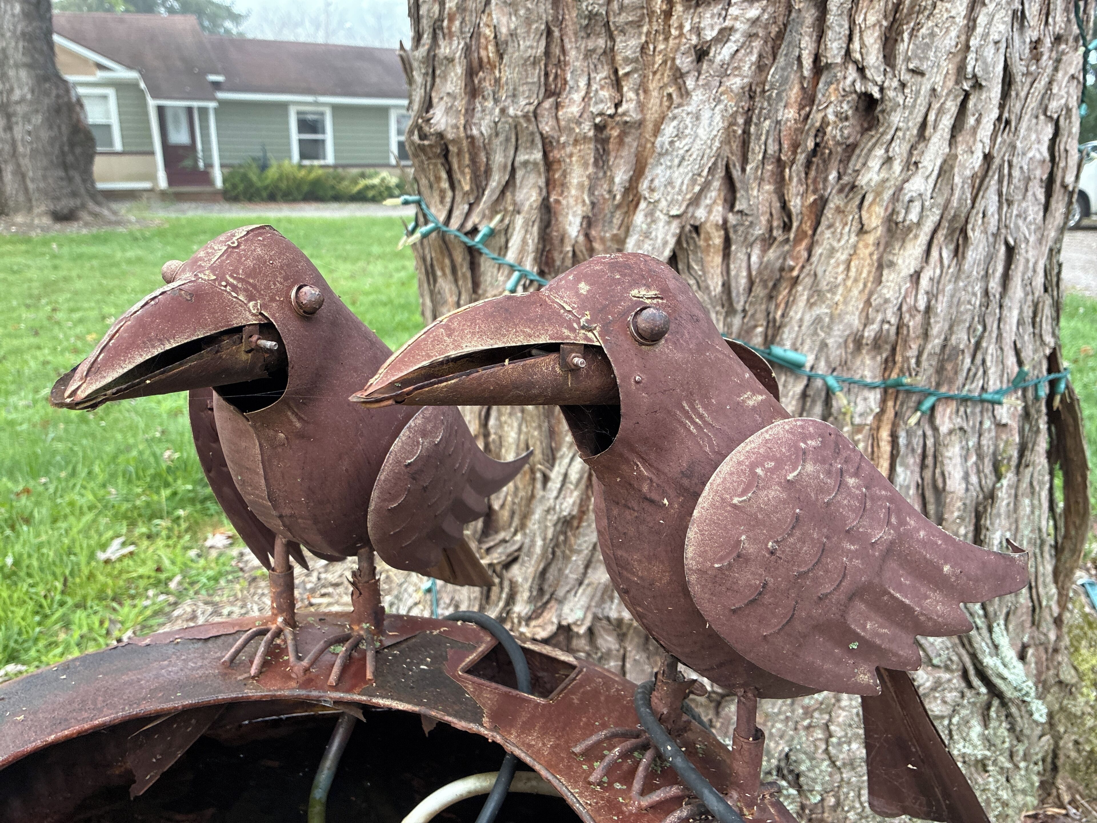 Two rusted metal bird sculptures mounted on a metal structure, with a tree and a house in the background.